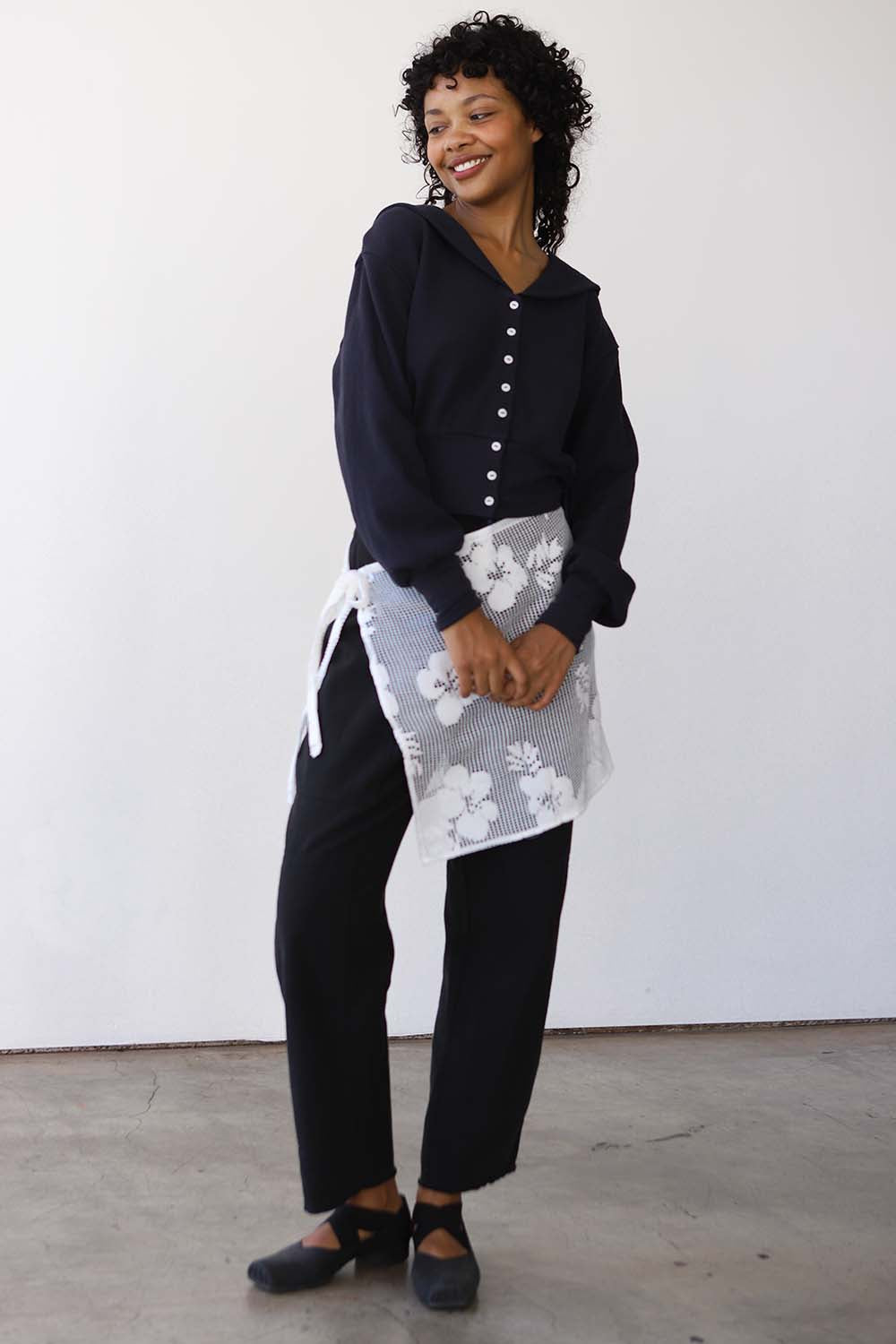 Woman wearing a black sweater and black pants with a white apron featuring black floral patterns against a white background.