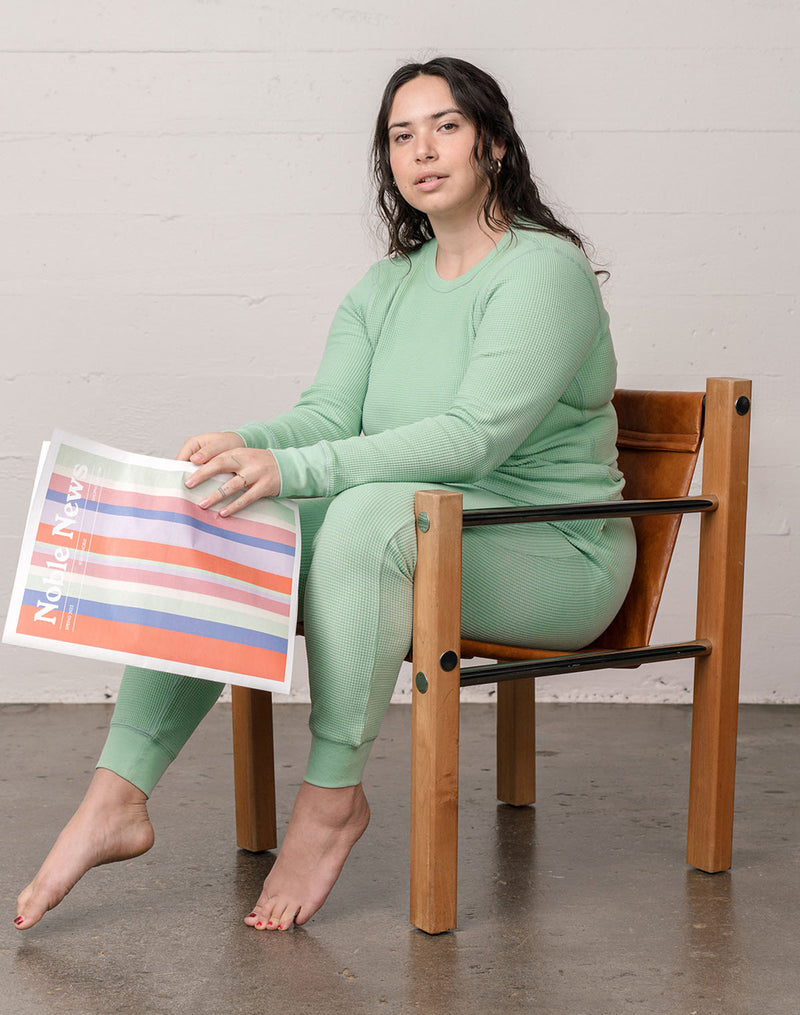 Woman wearing organic waffle thermal pajamas in sage reading a newspaper, sitting in a vintage modern chair. 