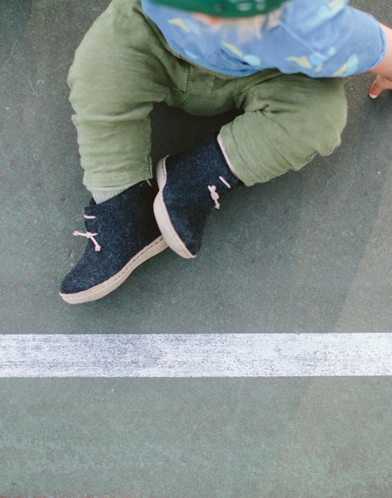 Baby sitting down wearing green pants and charcoal Glerups wool baby boots