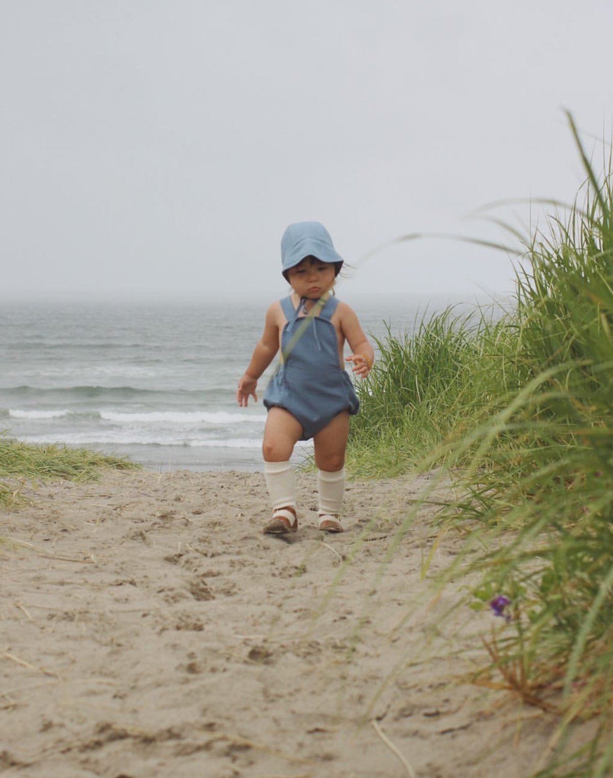 Baby walking on beach wearing a moon blue brimmed bonnet and sun suit with knee socks and sandals