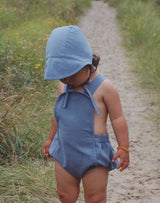 Baby walking on sand path wearing a moon blue brimmed bonnet and matching blue sun suit