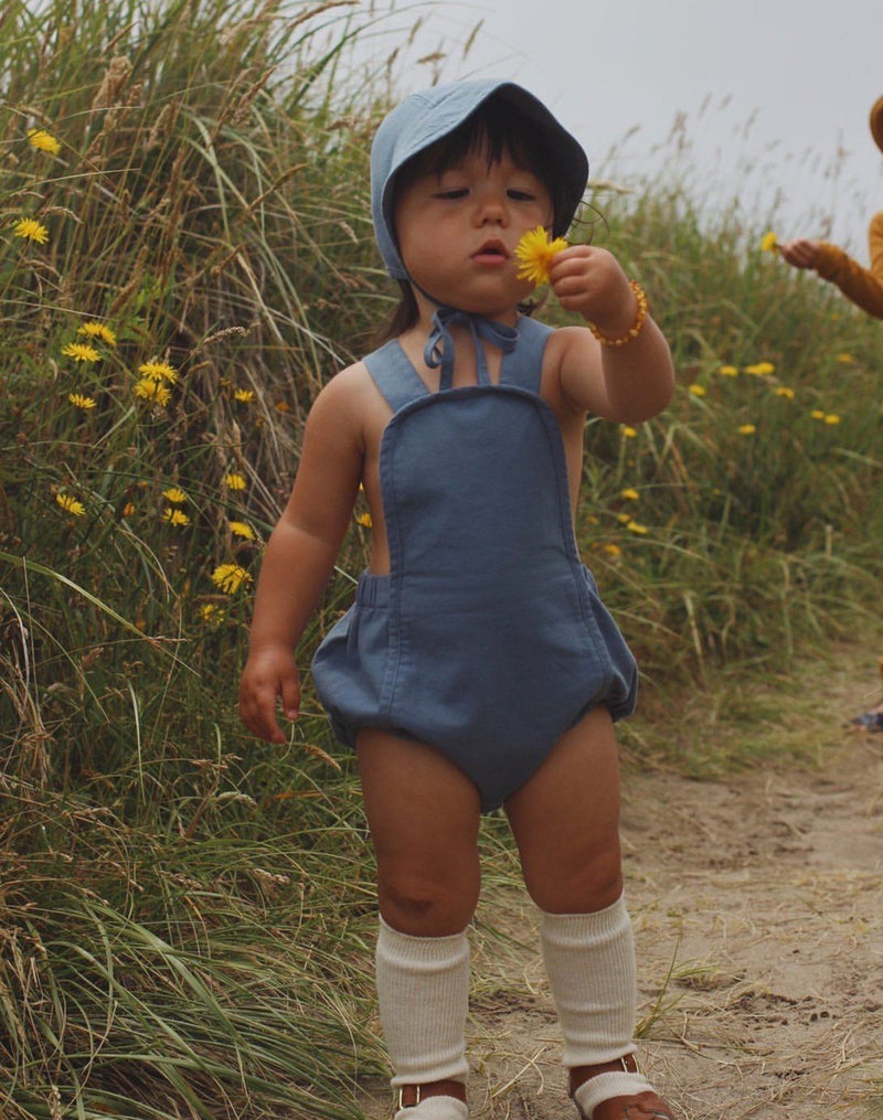 Baby picking yellow flowers in blue sunsuit and brimmed bonnet
