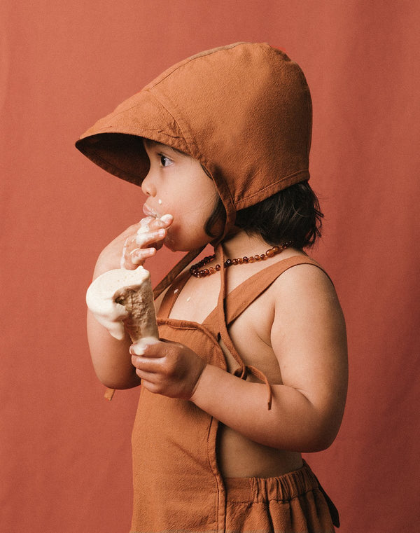 Baby licking an ice cream cone wearing a cinnamon colored brimmed bonnet and sun suit